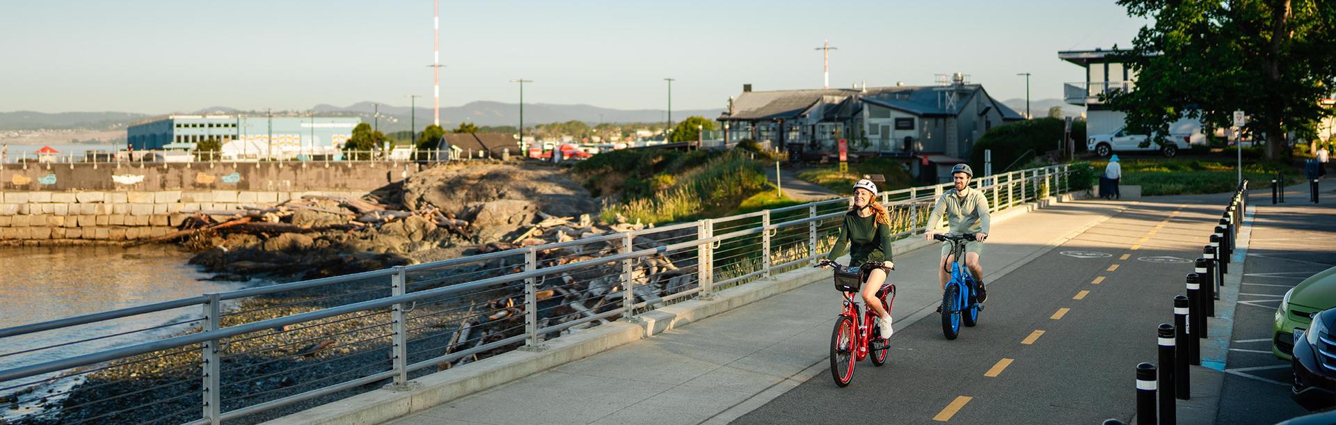A couple rides e-bikes along the Dallas Road walkway at sunrise in Victoria, BC