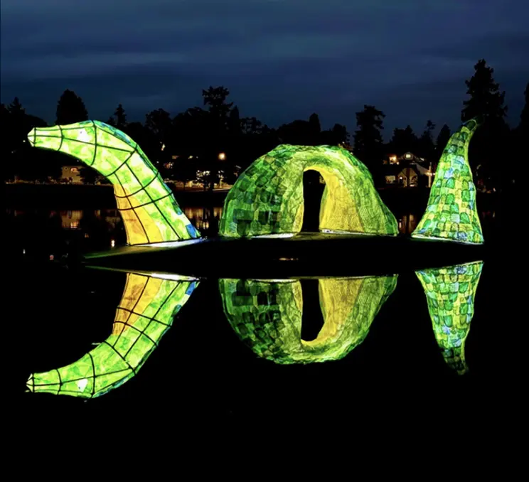 Illuminated green lantern sculpture reflected in calm water at night during the Esquimalt Lantern Festival.