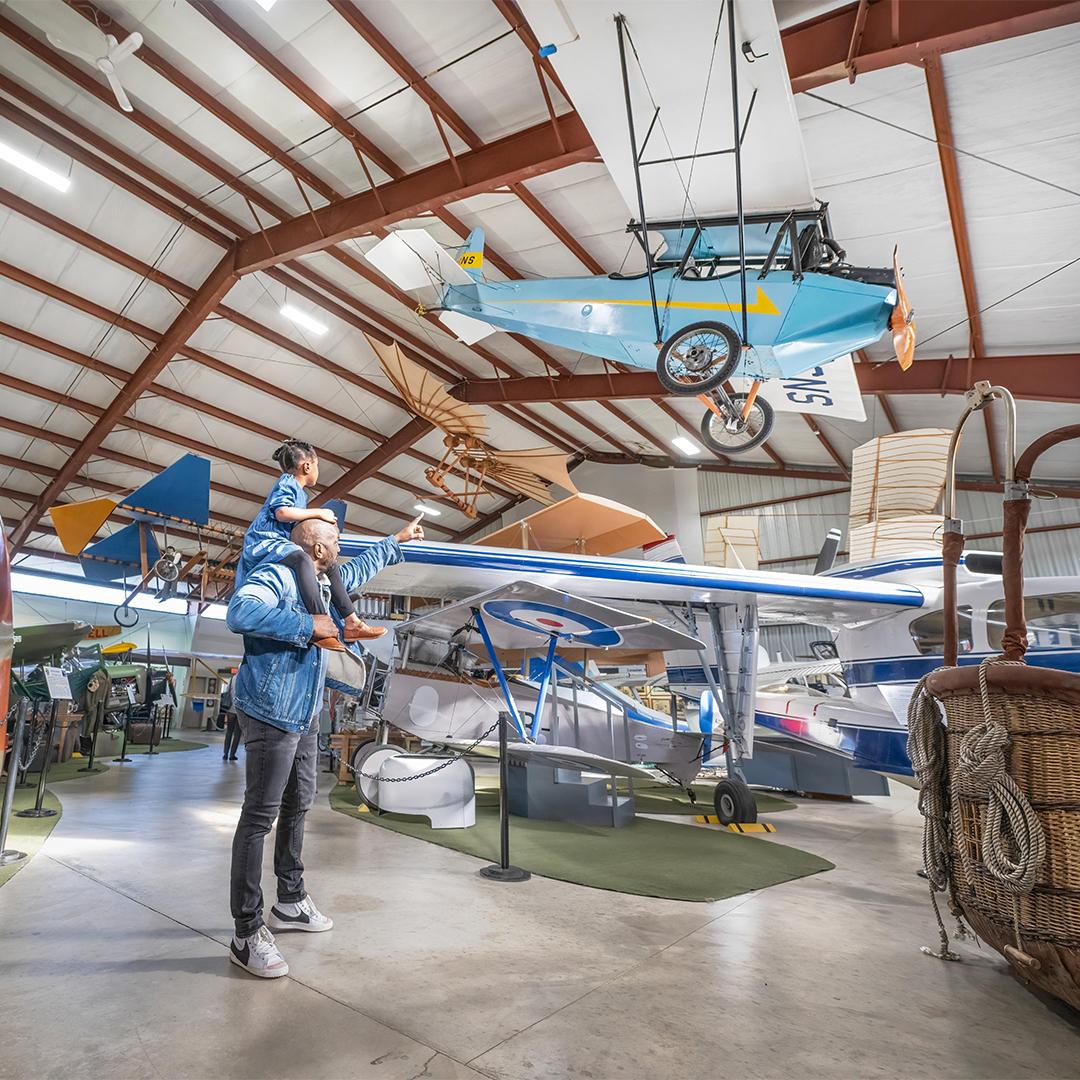 A father in a denim coat carries his daughter at the BC Aviation where a display of an aircraft is overhead, in Victoria, BC