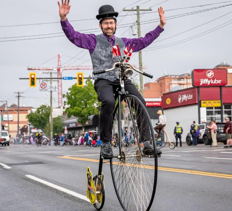 A performer dressed in purple and gray with a bowler hat rides a high wheel bicycle during the Victoria Day Parade in Victoria, BC