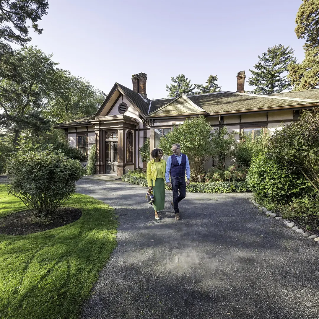 A woman in yellow and green and a man in blue walk down a pathway through the garden at Point Ellice House in Victoria, BC