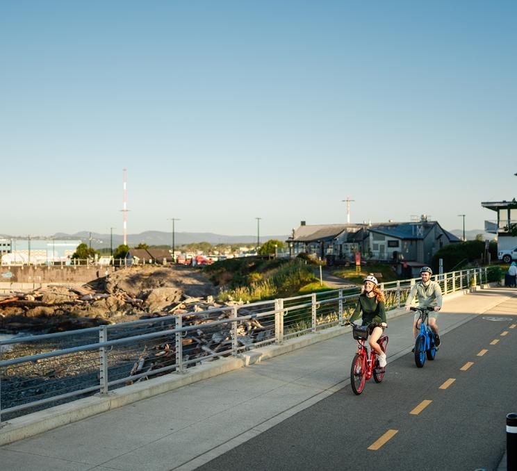 A couple rides e-bikes rented from Pedego along a Bike Path on Dallas Road in Victoria, BC