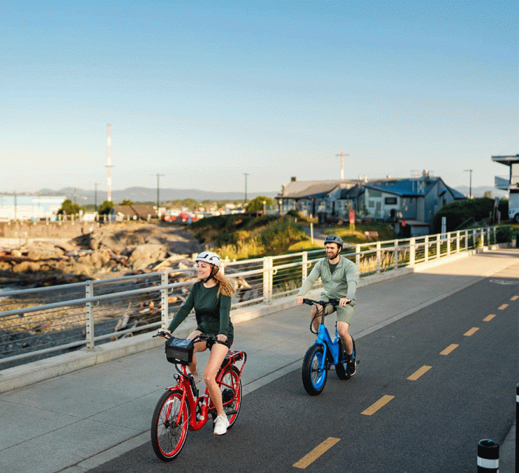 A couple rides red and blue e-bikes along a waterfront path on Dallas Road in Victoria, BC