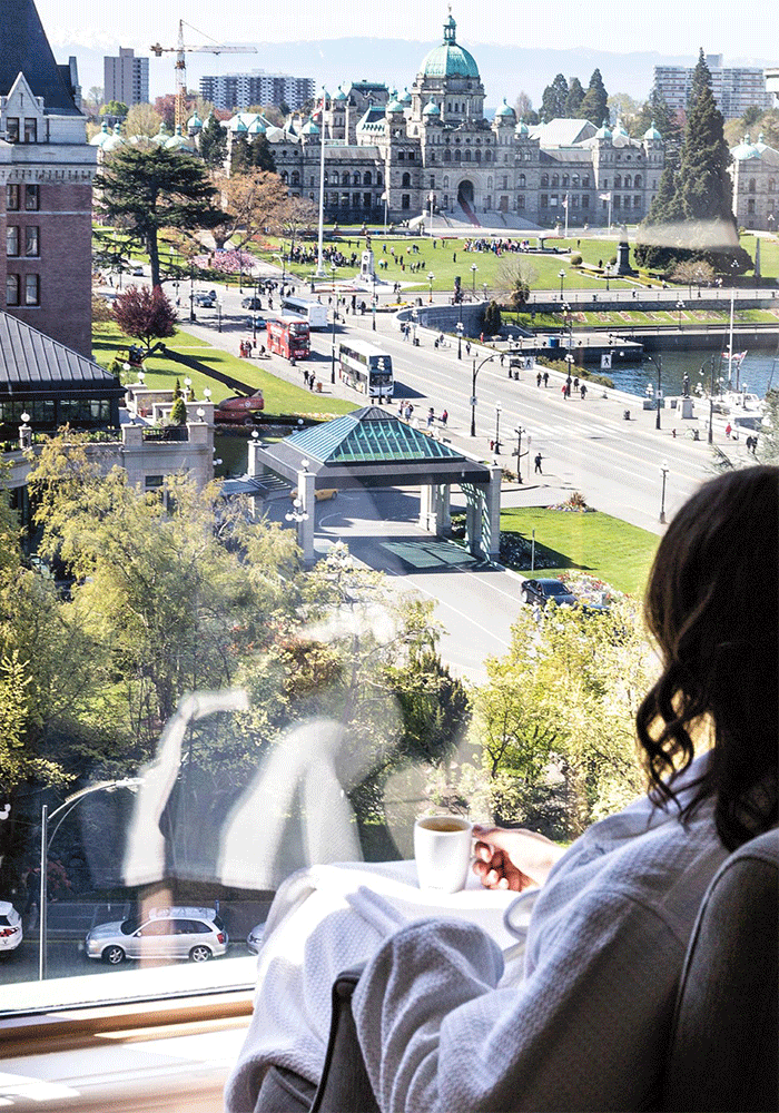 A woman in a robe enjoys a cup of coffee as she looks at toward the BC Parliament Buildings from the Magnolia Hotel & Spa in Victoria, BC