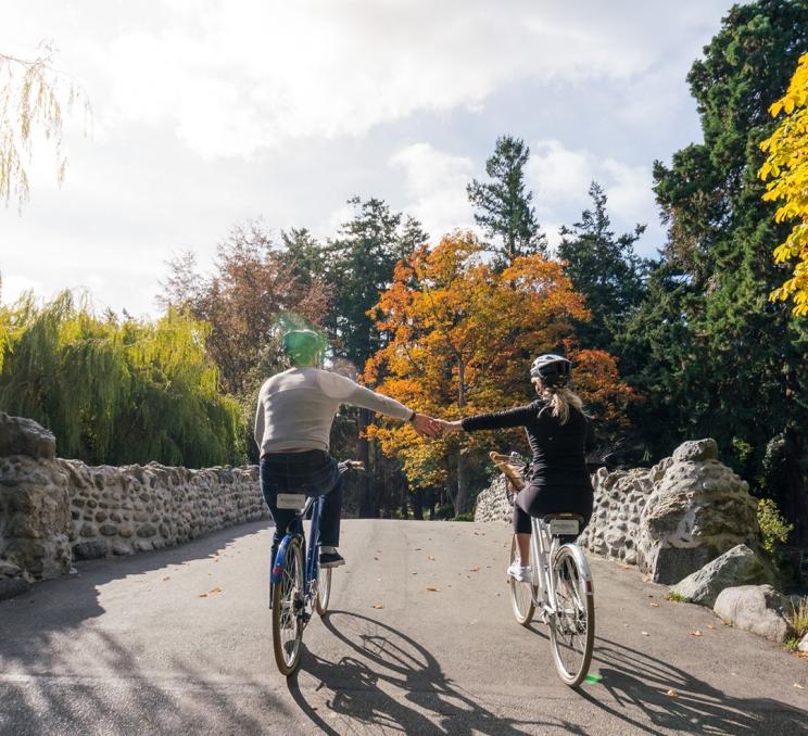 A couple ride bikes hand-in-hand over the stone bridge in Beacon Hill Park during the fall in Victoria, BC