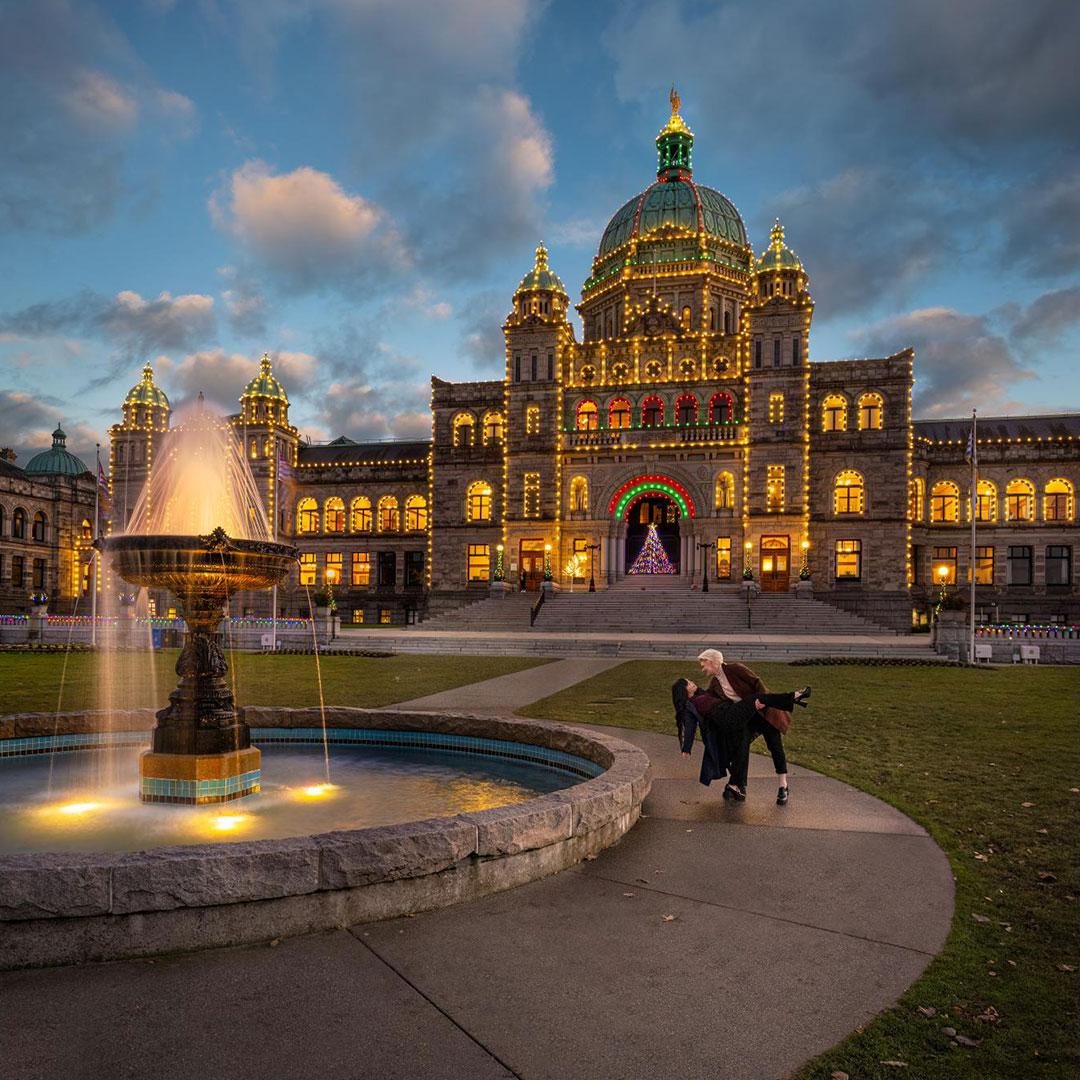 A female couple dances next to the fountain next the the fountain on the grounds of the BC Parliament Buildings, which are illuminated by holiday lights, in Victoria, BC