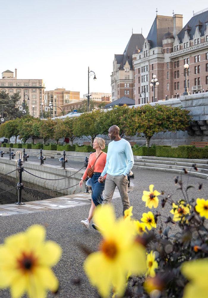 A couple walks hand-in-hand along the Lower Causeway of the Inner Harbour with the Fairmont Empress in the background in Victoria, BC