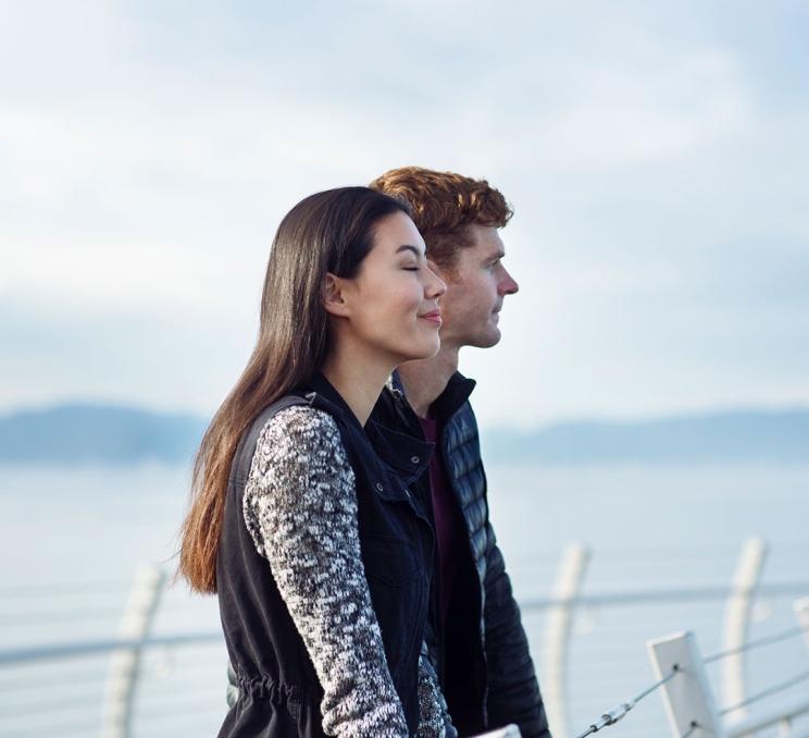 A couple stands along the railing of the Ogden Point Breakwater while looking out over the sea off the coast of Victoria, BC