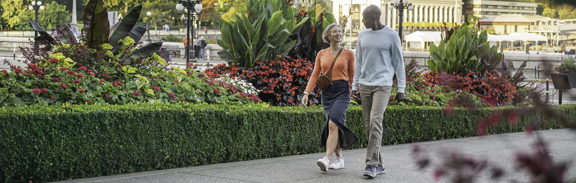 A couple walks hand-in-hand along a walkway past gardens in front of the Fairmont Empress Hotel on Victoria, BC's Inner Harbour