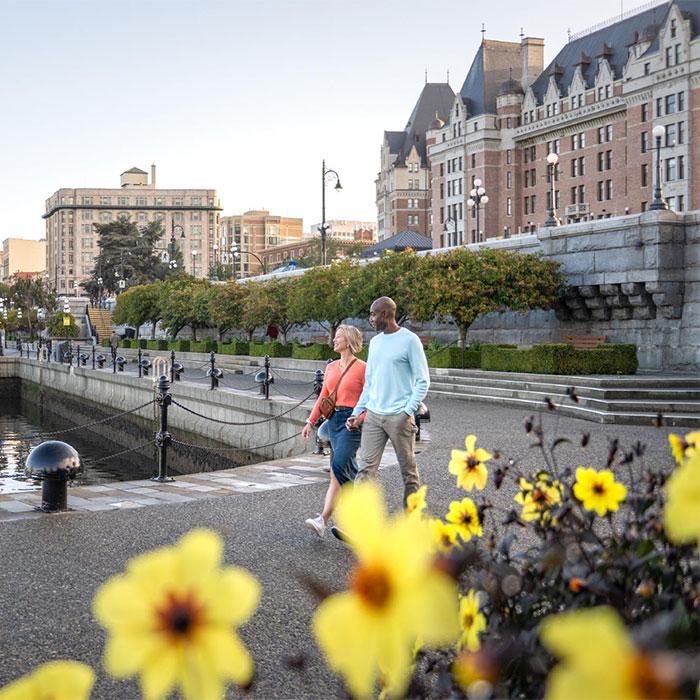 A couple walks hand-in-hand along the Lower Causeway of the Inner Harbour with the Fairmont Empress in the background in Victoria, BC