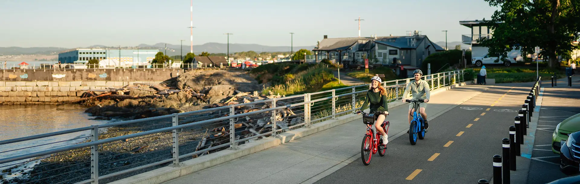 A couple ride red and blue e-bikes along the bike path on Dallas Road in Victoria, BC's James Bay neighbourhood