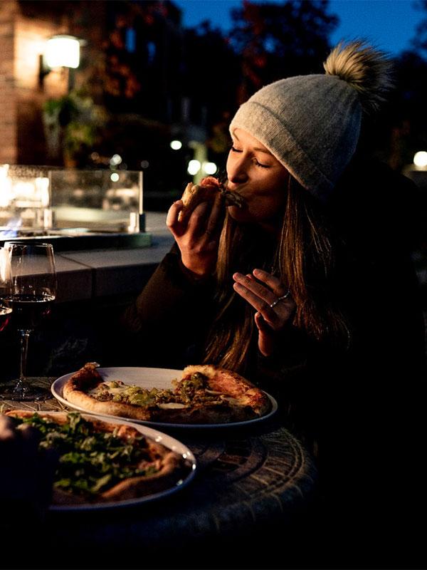 A woman wearing a toque bites into a slice of pizza while dining on the patio during the evening at FARO Handcrafted Pizza & Tasting Room in Victoria, BC