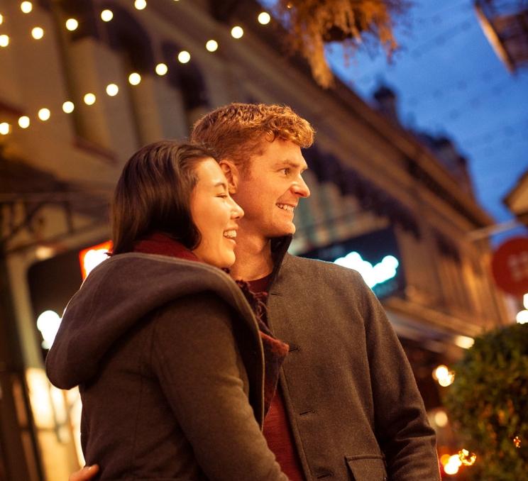 A couple in Trounce Alley stands beneath twinkling lights at dusk in Victoria, BC
