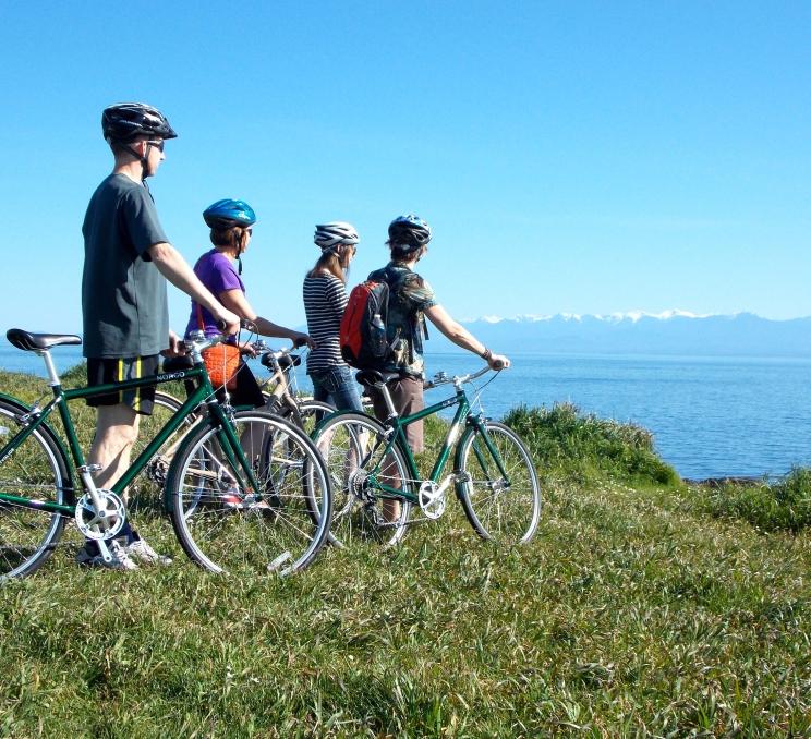 A family biking along Dallas Road in Victoria, BC, with ocean views and the Olympic Mountains in the distance.