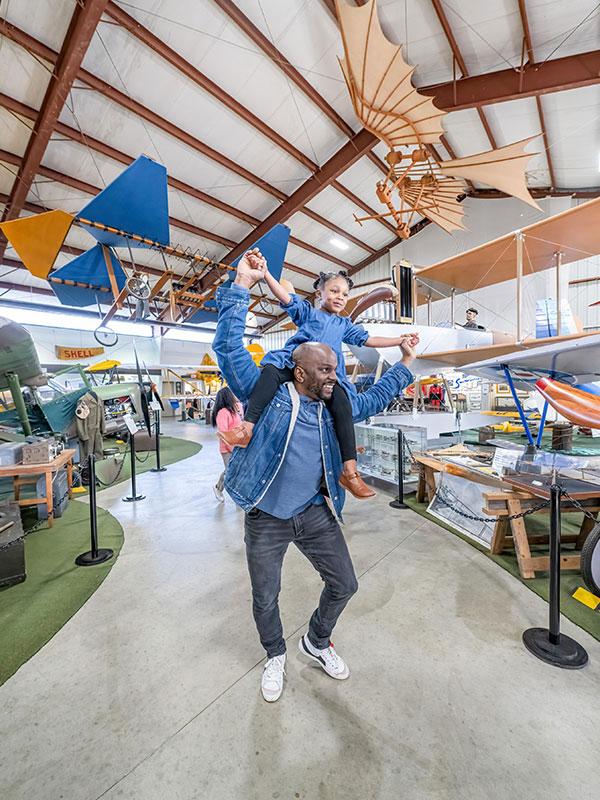 Parent lifting child on shoulders inside the BC Aviation Museum in Victoria BC surrounded by historic aircraft and early flight exhibits