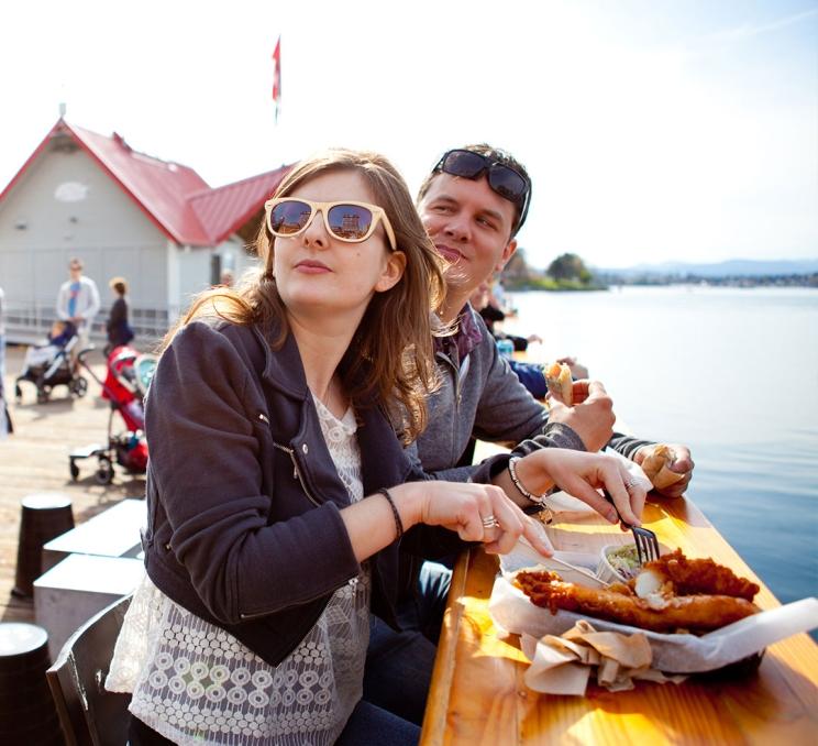 A couple enjoys dockside fish & chips outside of Red Fish Blue Fish on Victoria, BC's Inner Harbour