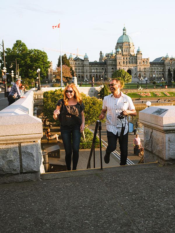 A couple walks up a flight of stairs from the Lower Causeway to the Upper Causeway of Victoria, BC's Inner Harbour at sunset