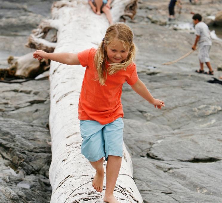 Child balancing on a large driftwood log at a rocky beach in Greater Victoria, BC with ocean and mountains in the background