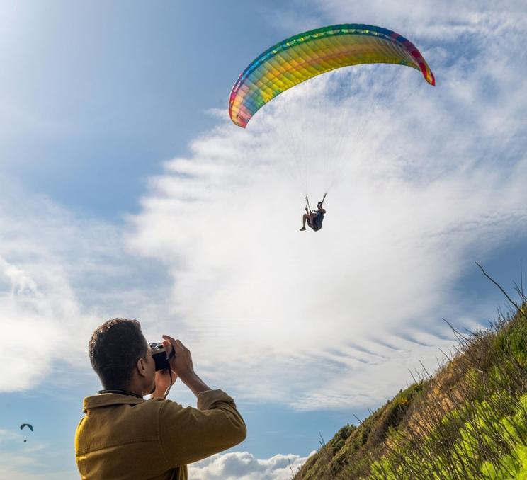 A person takes a photo of a paraglider flying overhead at Clover Point along the Dallas Road water font in Victoria, BC