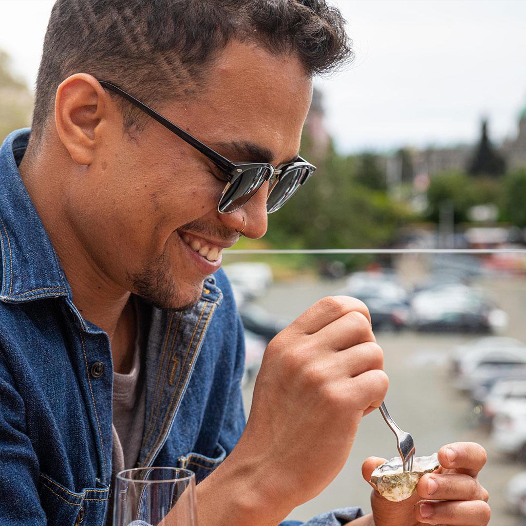 A visitor to Victoria, BC digs into an oyster on the half shell while sitting on the patio at Finn's Seafood, Chops & Cocktails on the Inner Harbour