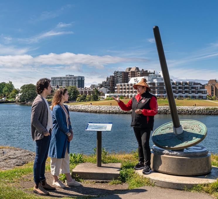 A couple watches as a tour guide from Explore Songhees speaks of the history of the 7 Signs of the Lekwungen sculpture behind her in the Vic West neighbourhood of Victoria, BC