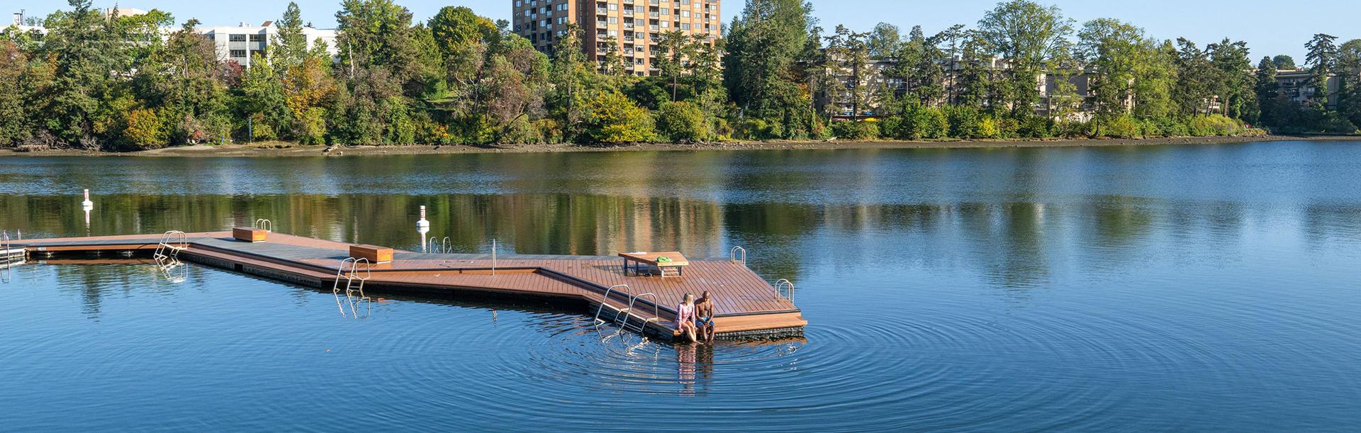 A couple dips there feet in the water off the edge of the dock at Banfield Park in the Vic West neighbourhood of Victoria, BC