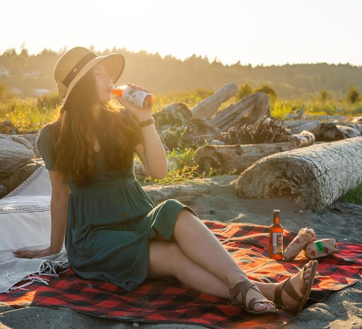 A woman drinking kombucha during a picnic while sat on a beach in Greater Victoria, BC