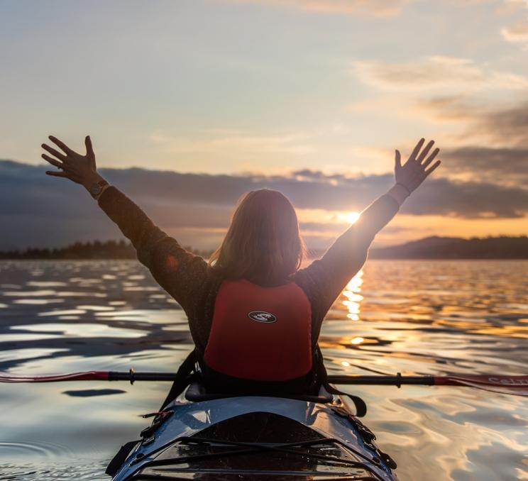 A woman throws up her hands while kayaking at sunset in Victoria, BC
