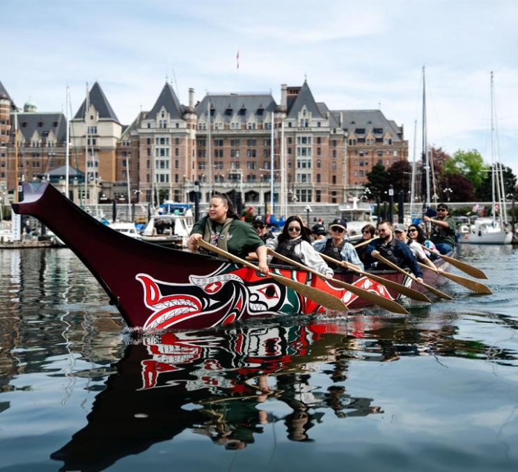 Participants paddle a traditional Indigenous canoe through Victoria's Inner Harbour with the Fairmont Empress and marina visible in the background.