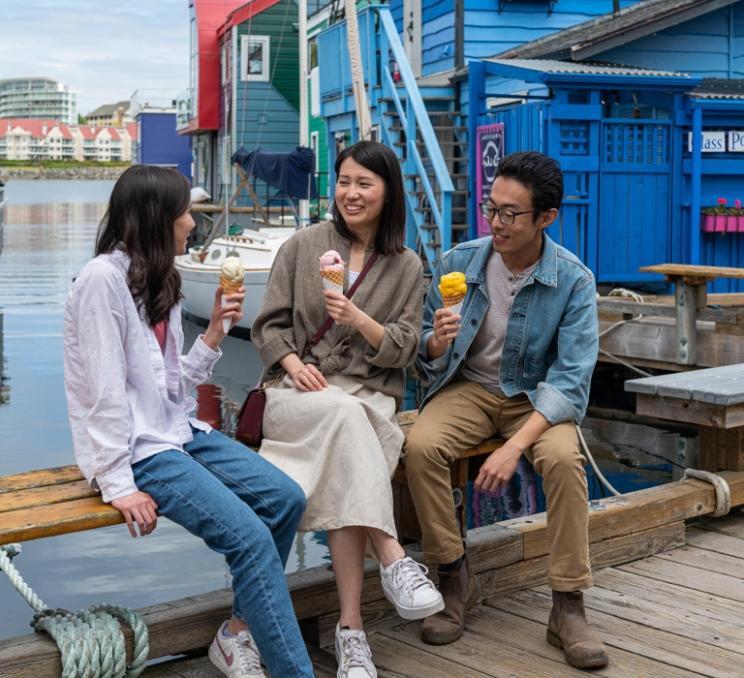 Three people enjoy ice cream while sitting on a dock at Fisherman's Wharf in Victoria, surrounded by colourful floating homes and boats.