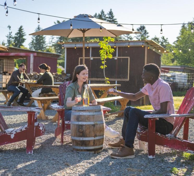 Visitors enjoy drinks and cider in an outdoor seating area at Junction Orchard & Cidery in Saanich on a sunny summer afternoon.
