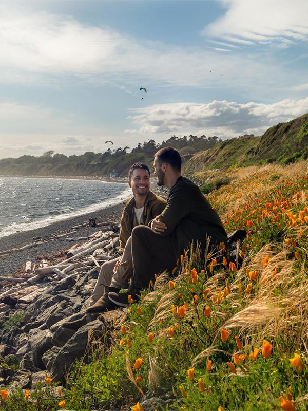 A male couple enjoy ocean views while sitting amongst the the wildflowers at Clover Point along the Dallas Road coastline in Victoria, BC