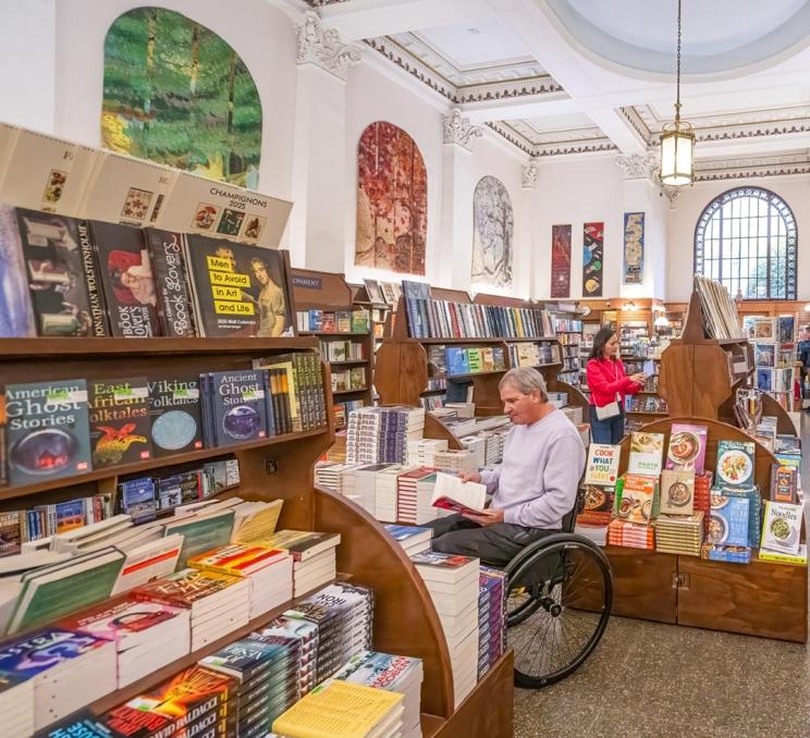 A traveller with accessible needs who uses a wheelchair browses the books in Munro's Books in Victoria, BC