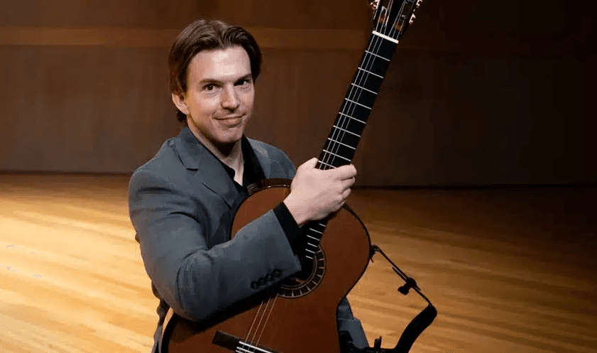 Person seated on a wooden stage holding a classical guitar.