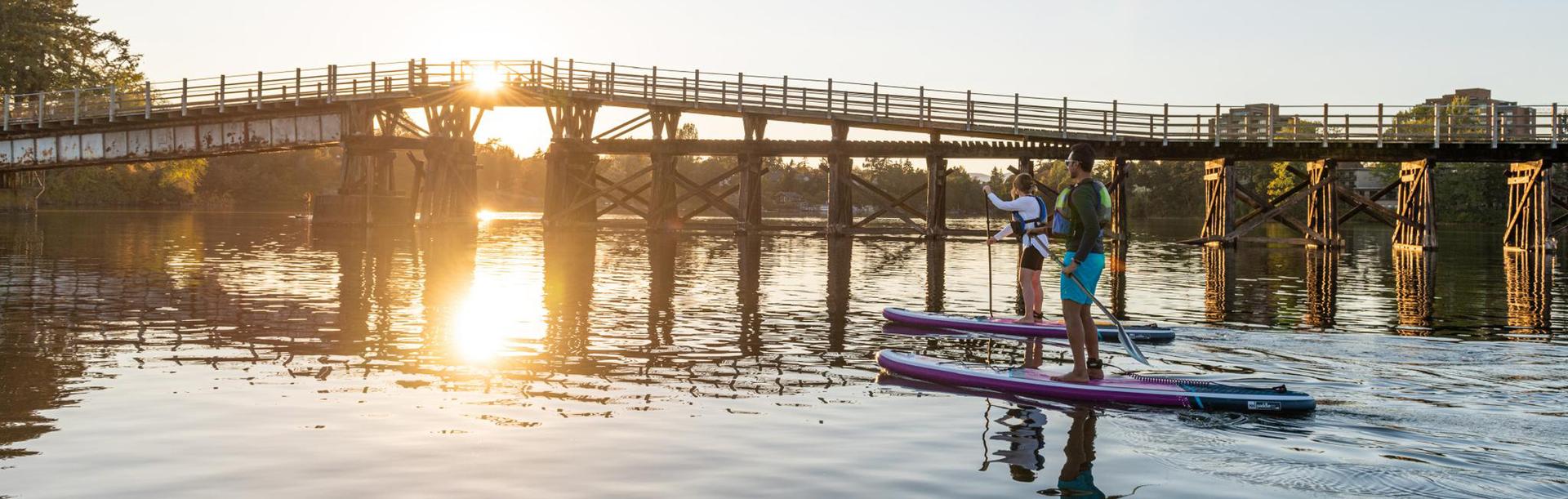 Two people paddleboard beneath the historic Selkirk Trestle spanning the Gorge Waterway in Victoria during a golden summer sunset.