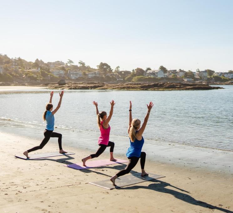 Three people practicing yoga on Gonzales Beach beside calm water on a sunny morning in Victoria, BC.