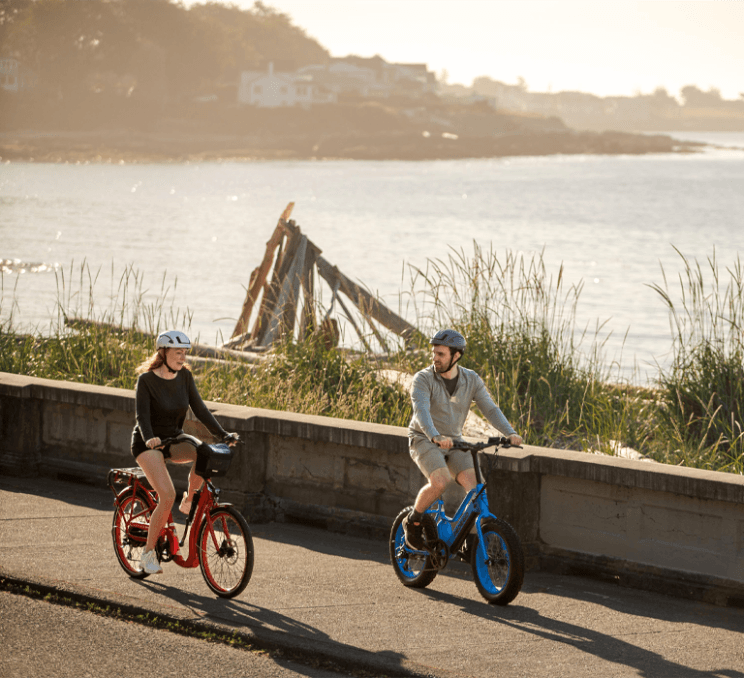 A couple rides red and blue e-bikes along a waterfront path on Dallas Road in Victoria, BC