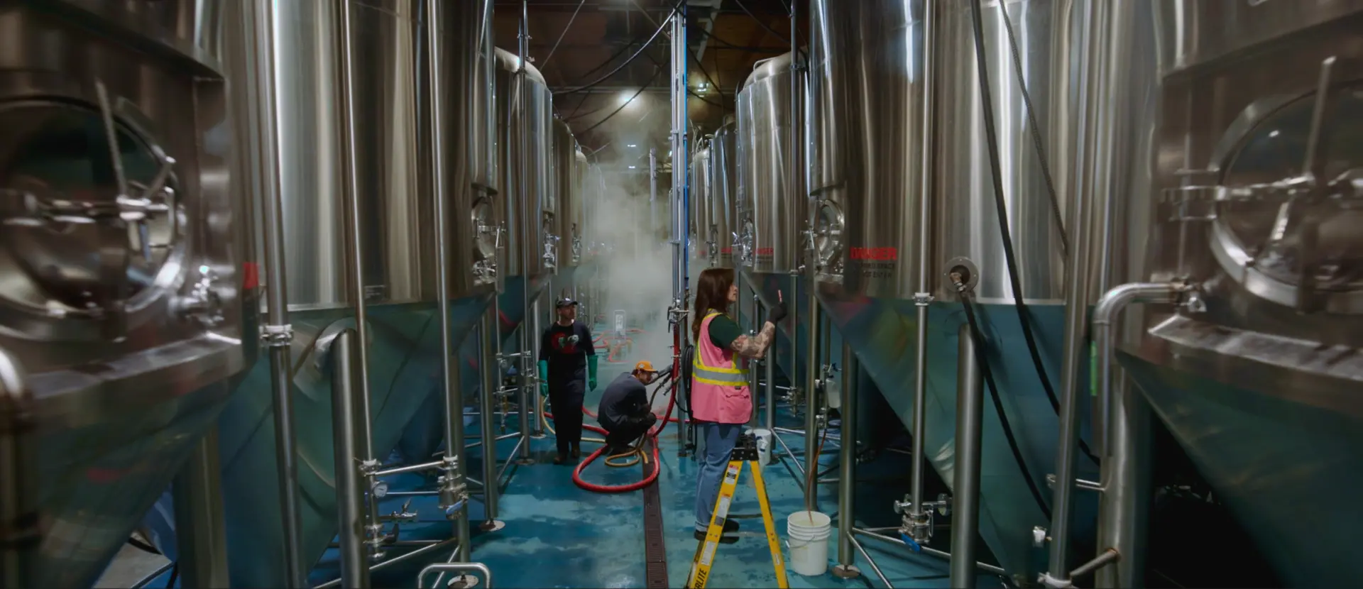 Three brewery workers tend equipment among rows of large stainless-steel fermentation tanks.