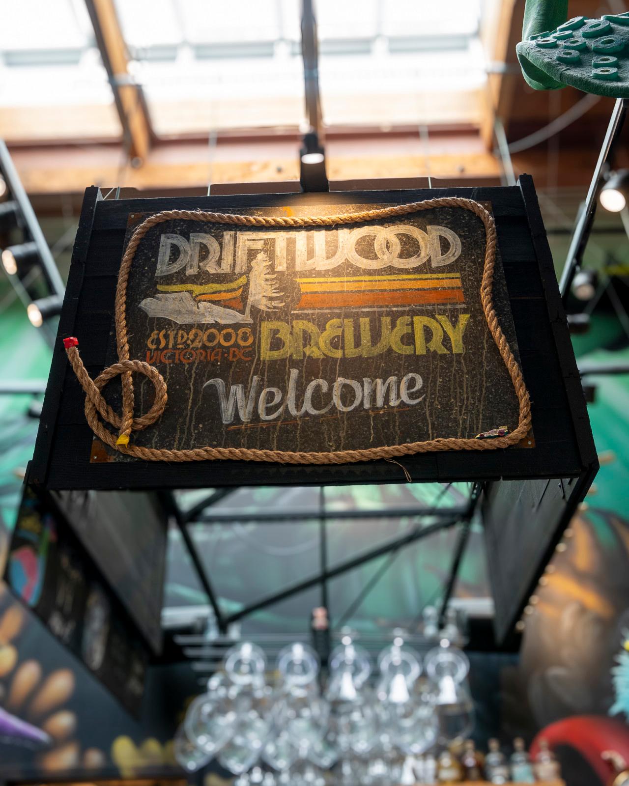 Vintage-style Driftwood Brewery welcome sign framed with rope and suspended above the taproom bar, with hanging glassware visible below.
