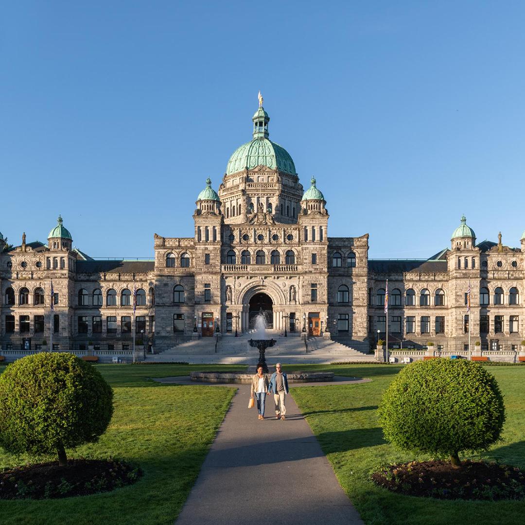 A couple walks hand-in-hand along a pathway in front of the BC Parliament Buildings in Victoria, BC
